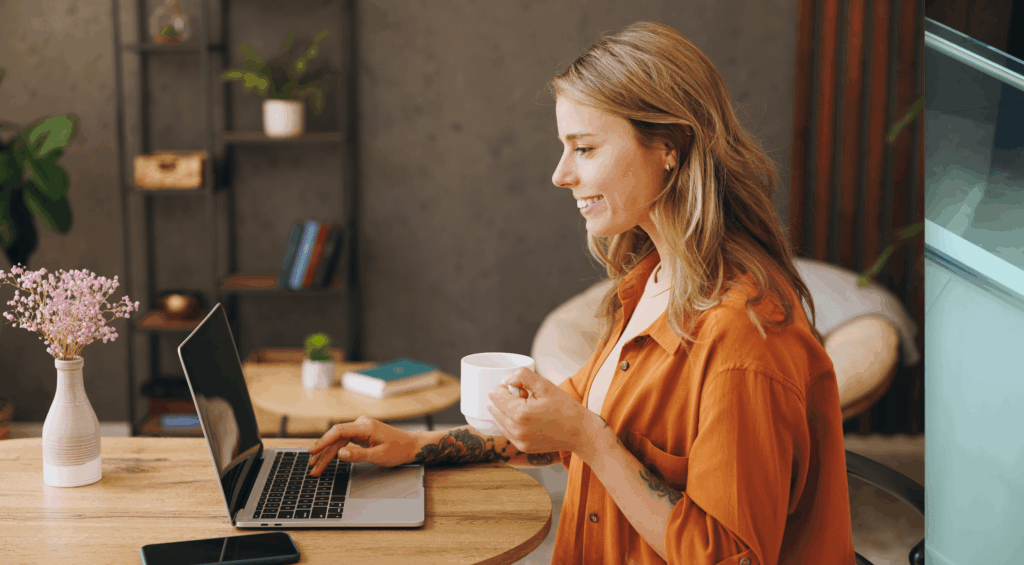 A young woman networking online on her laptop, whilst drinking from a mug.