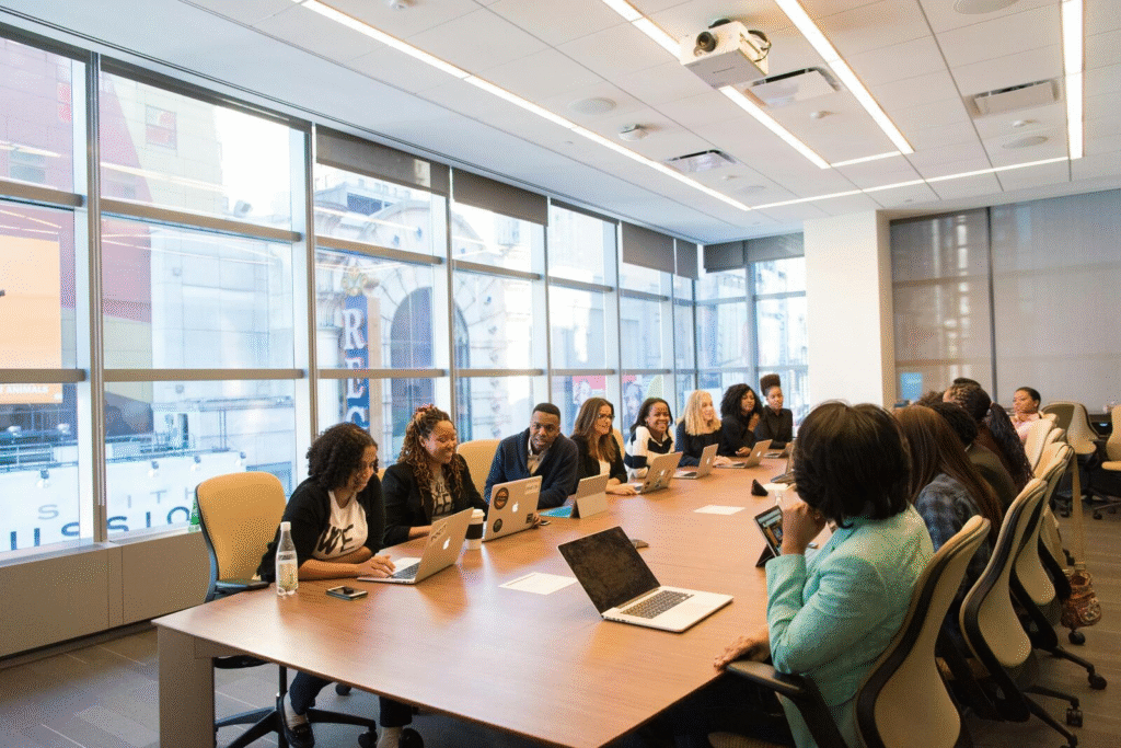 Large conference room with a group meeting around a long boardroom table, working on laptops beside floor-to-ceiling windows and a ceiling-mounted projector.
