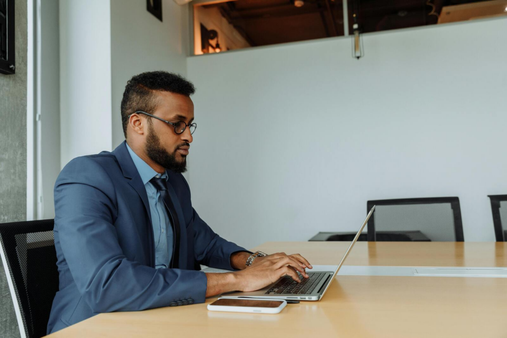 Man in a blue suit working on a laptop at a conference table, with a smartphone beside him in a modern office.