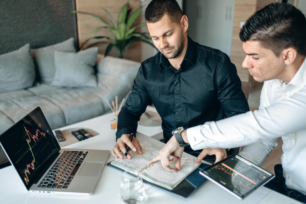 Two men reviewing notes at a table with a laptop and tablet displaying financial charts, suggesting an investor meeting or funding discussion.