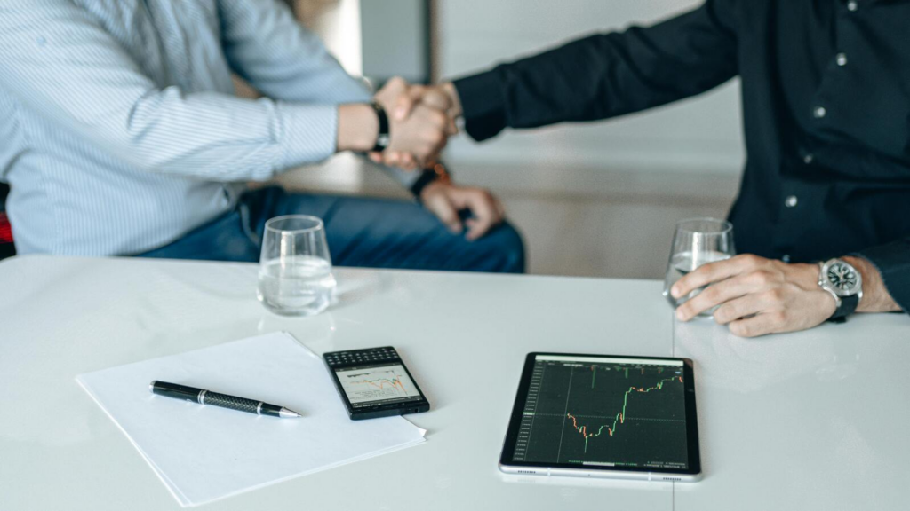 Two people shaking hands across a table with a tablet and phone showing market charts, alongside a pen, paper, and glasses of water.